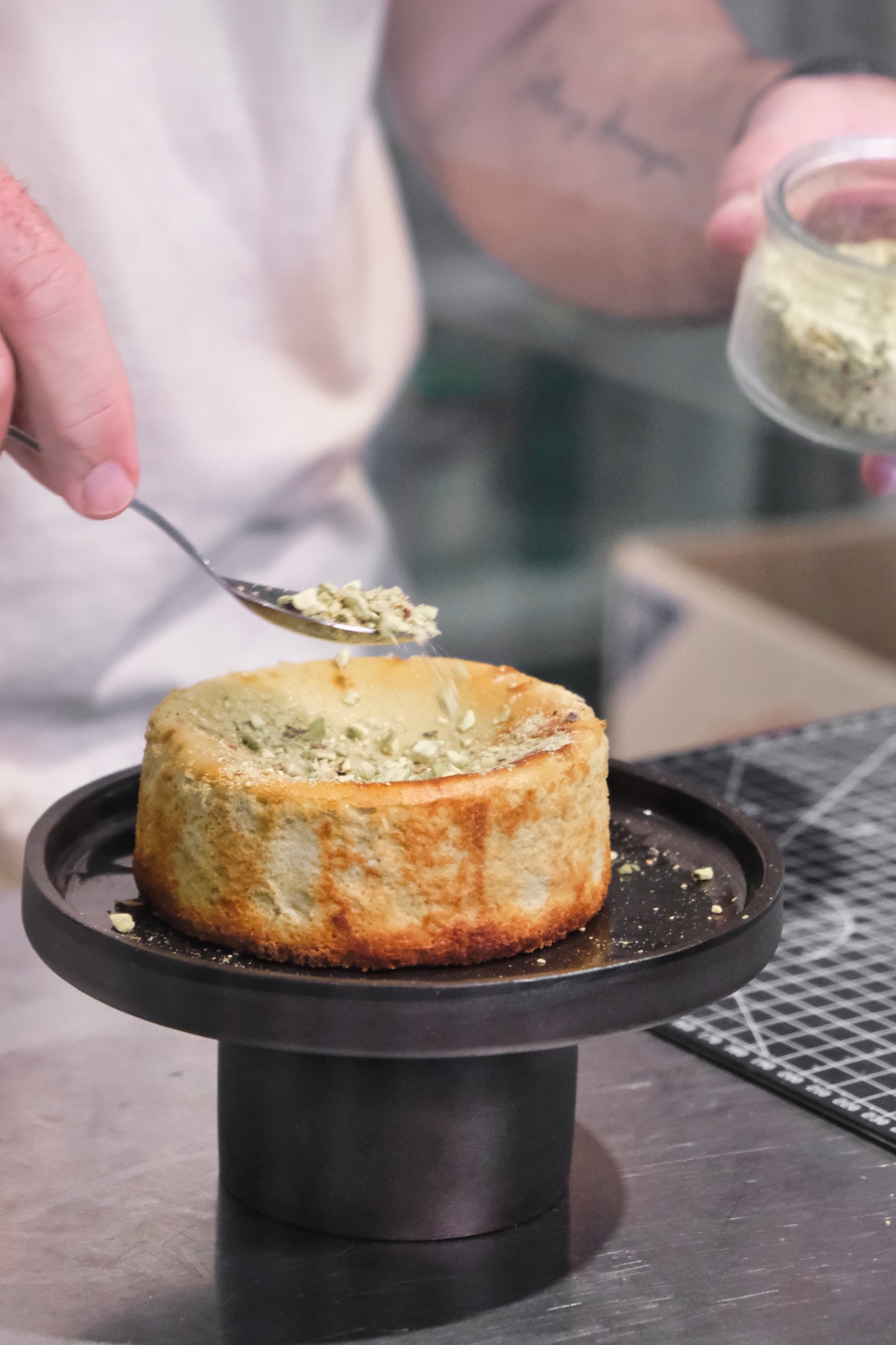 Manos espolvoreando pistacho picado sobre una tarta vasca, preparación artesanal